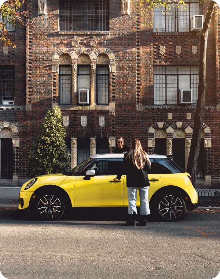 Two people enter a MINI Cooper 4 Door in front of a brick building.